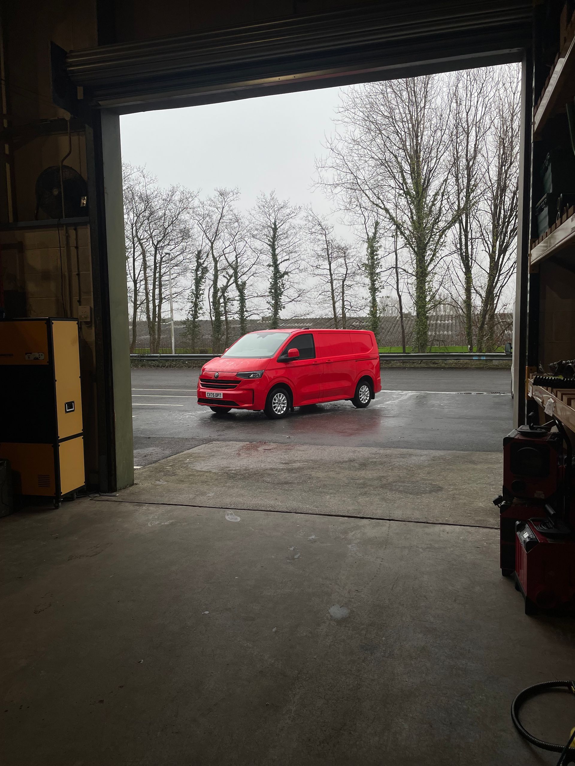 A red van is parked on wet asphalt outside a garage, viewed through an open bay door.