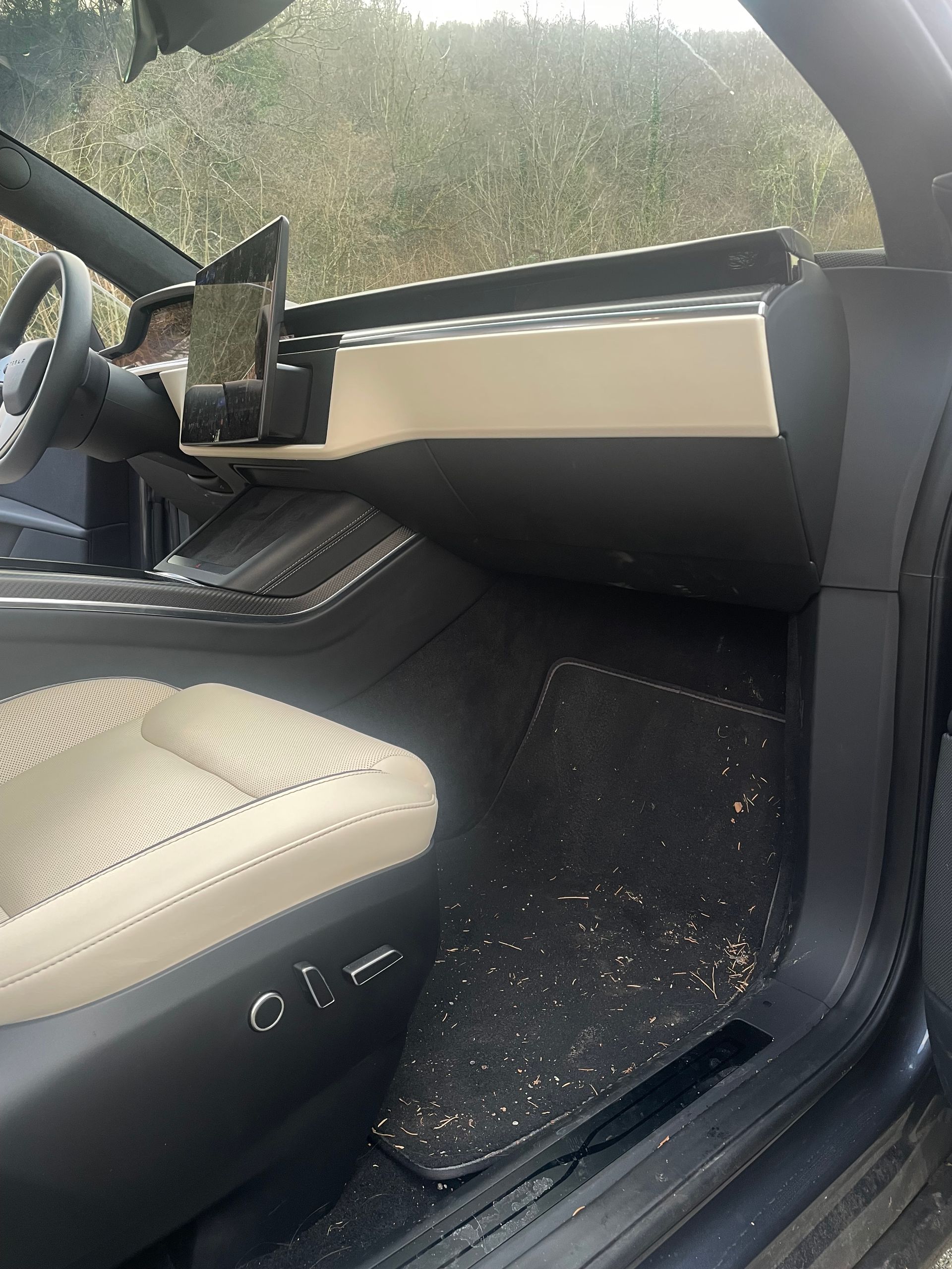 Interior of a car with a black and beige dashboard, beige seat, and debris scattered across the floor mat.