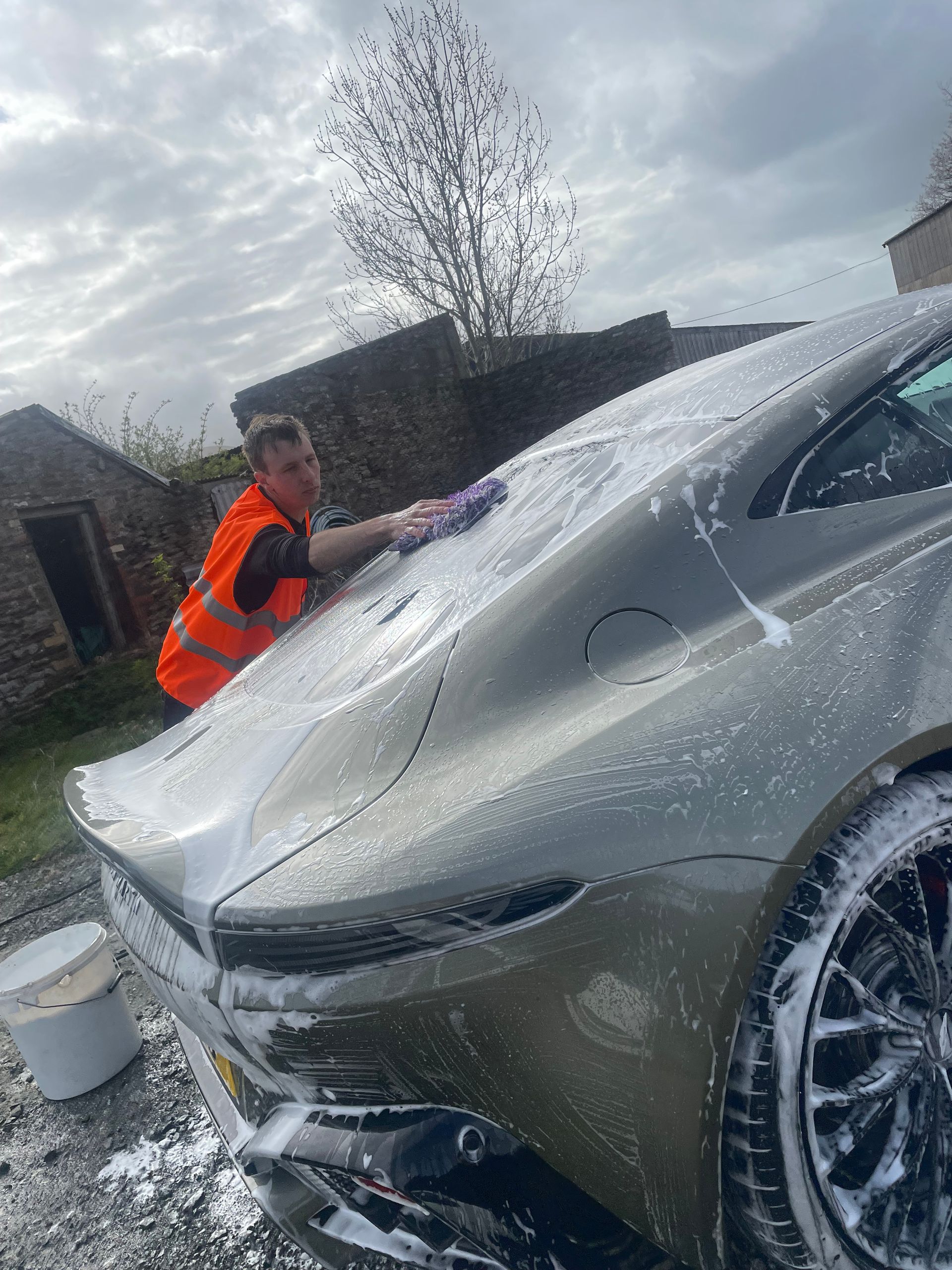 A person in a high-visibility orange vest washes a silver sports car covered in soap suds outdoors.