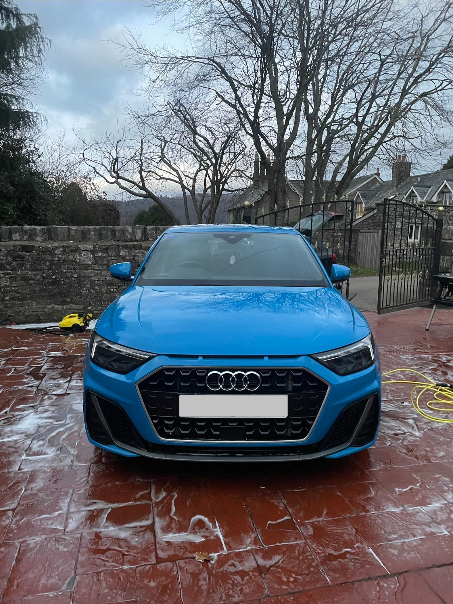 A bright blue Audi hatchback parked on a paved driveway covered in soapy water, viewed from the front.