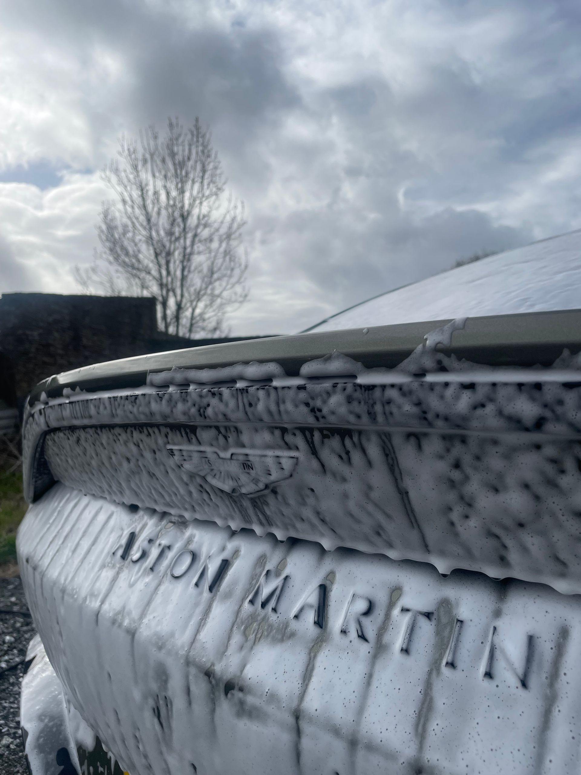 The rear of an Aston Martin car covered in thick, white soap suds under a cloudy sky.