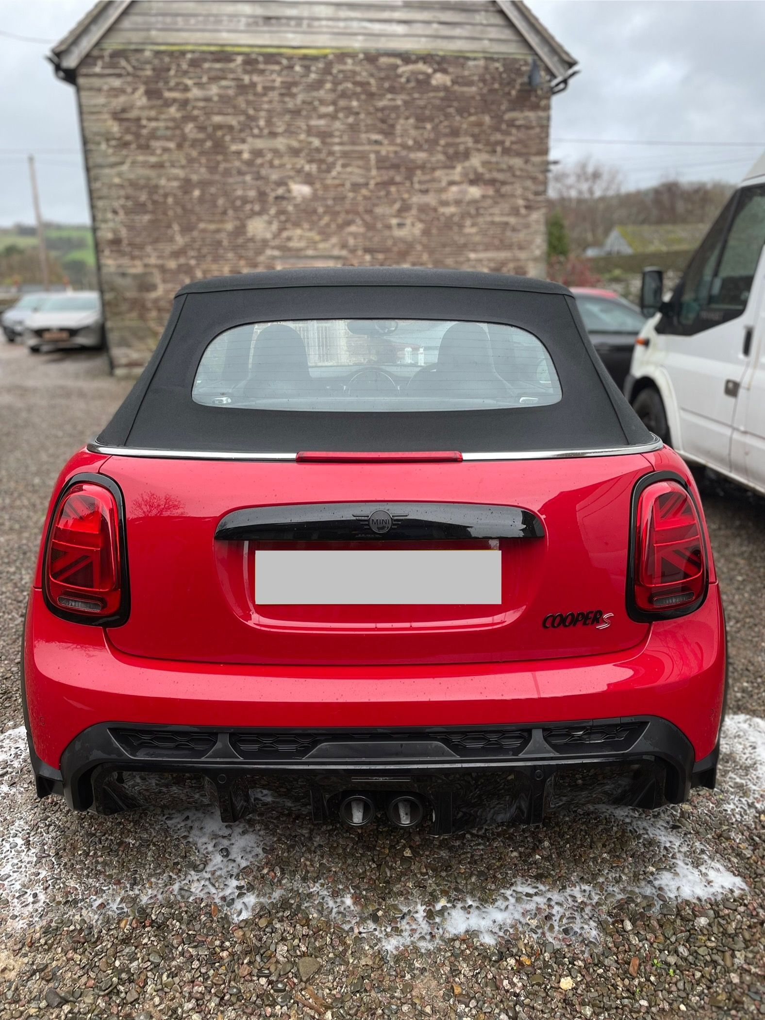 A red Mini Cooper convertible seen from the rear, parked on a gravel lot in front of a stone building.