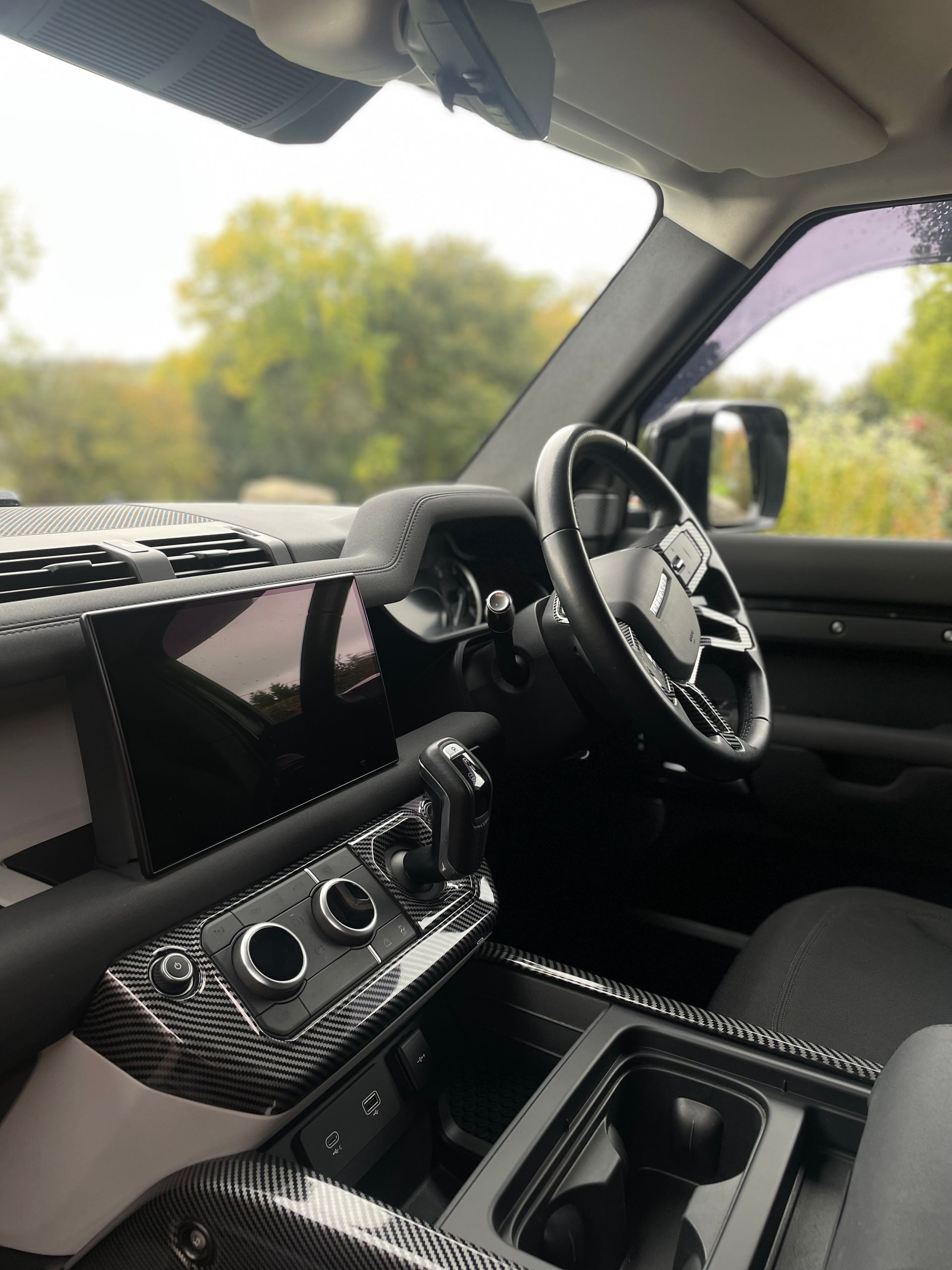 Interior view of a modern vehicle dashboard and steering wheel featuring carbon fiber trim and a large central screen.