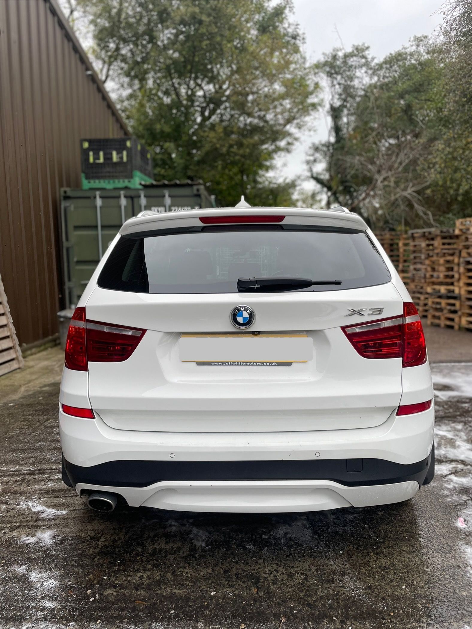 A white BMW X3 SUV parked on a paved outdoor lot with trees and a building in the background.