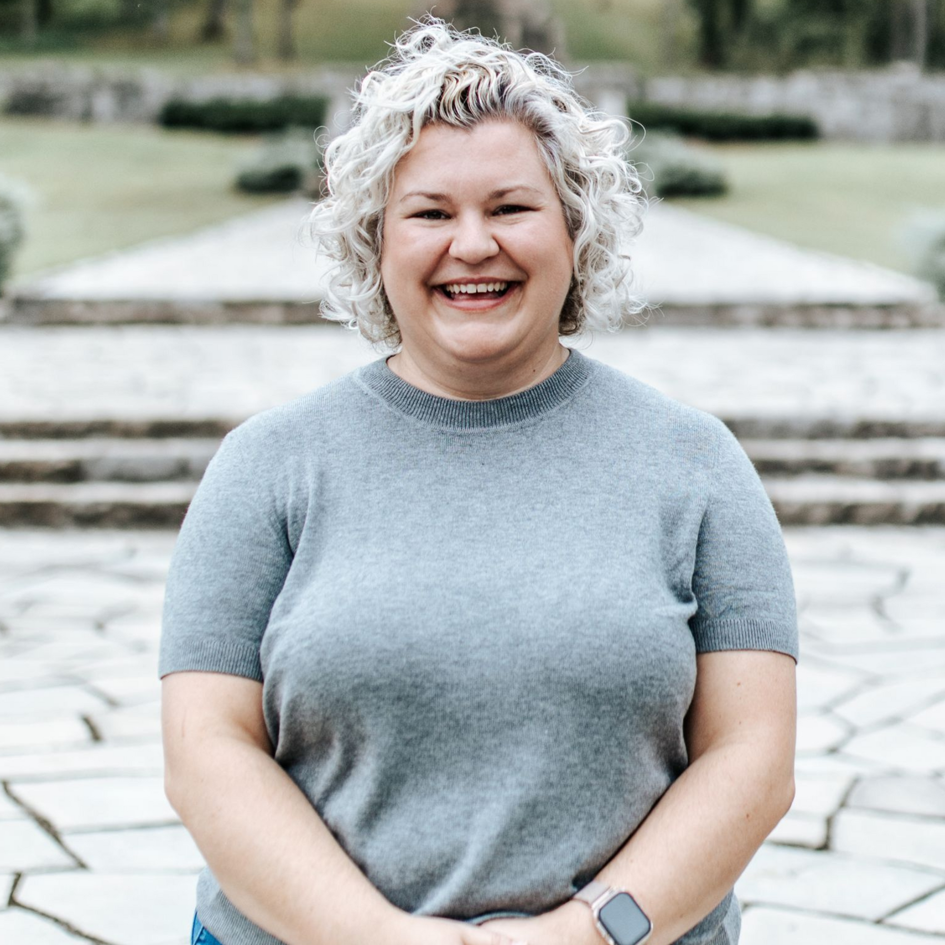 Woman with curly gray hair smiles outdoors, wearing a gray shirt, with a stone background.