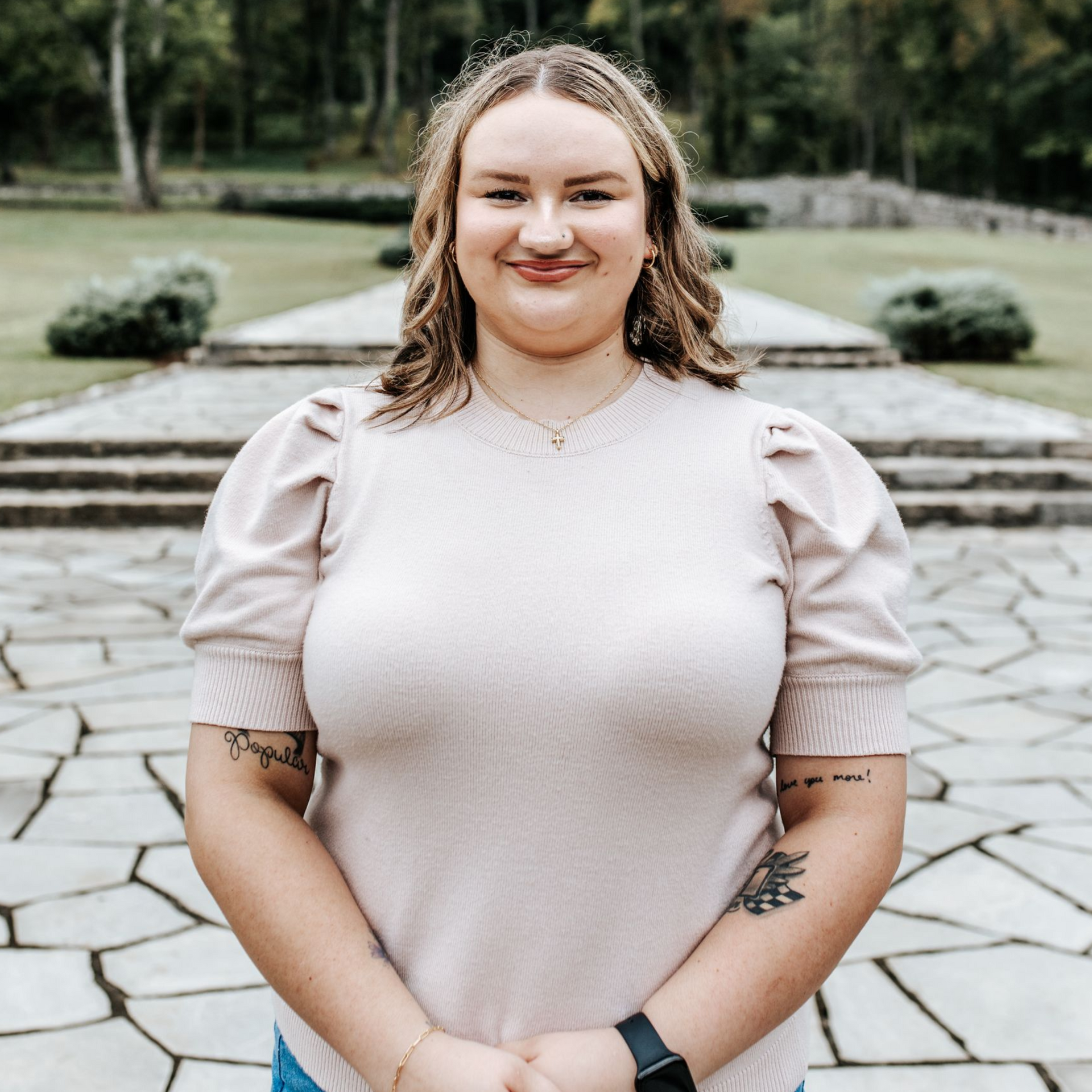 Woman in a light pink top smiles outdoors, with trees in the background. She has tattoos and a smartwatch.