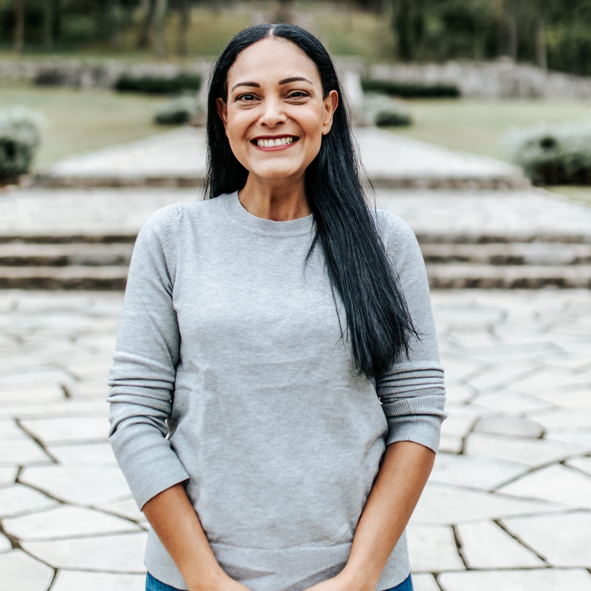 Woman with dark hair smiles, wearing a grey sweater, standing outdoors. Stone steps in background.