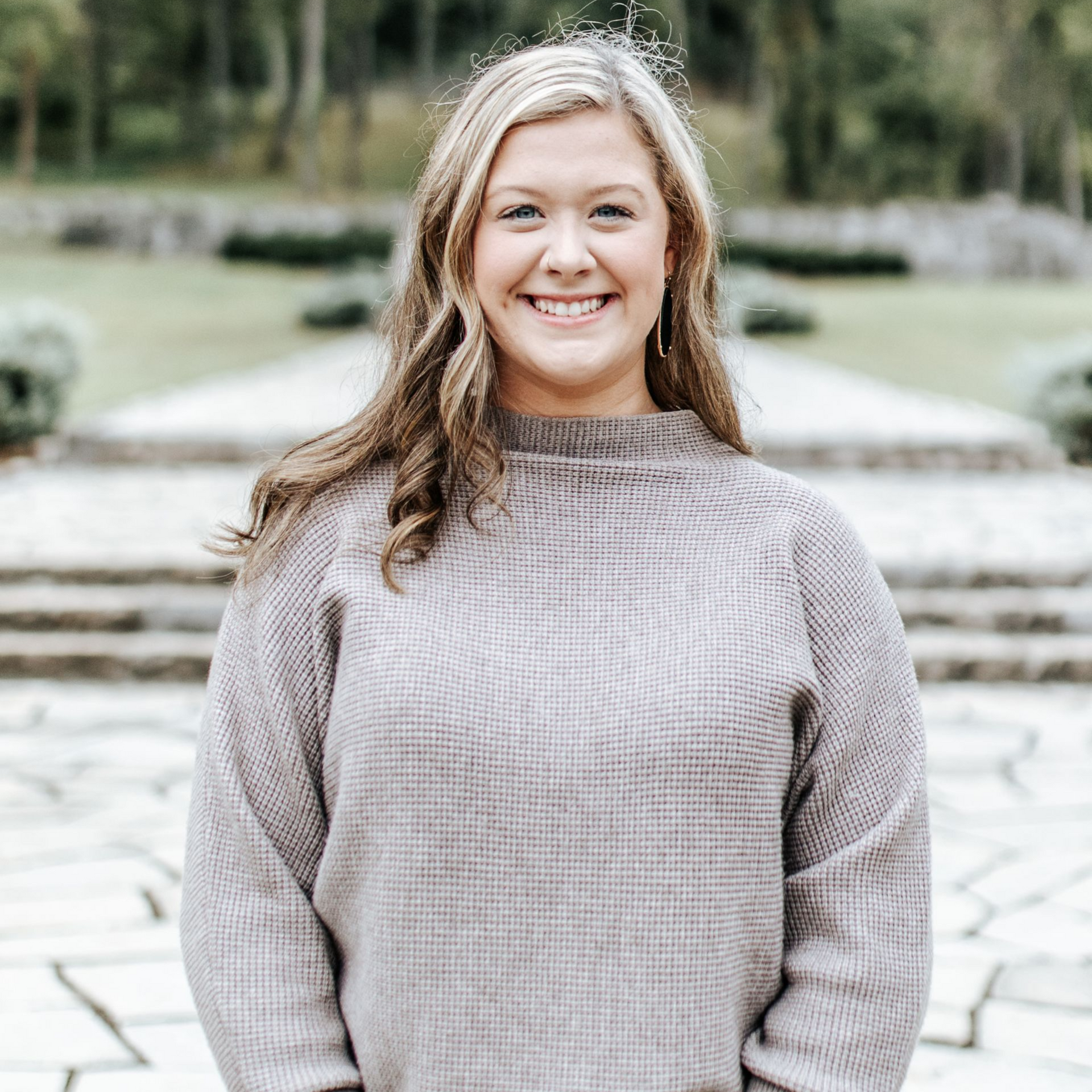 Woman smiling outdoors, wearing a tan sweater. Background includes stone steps and trees.