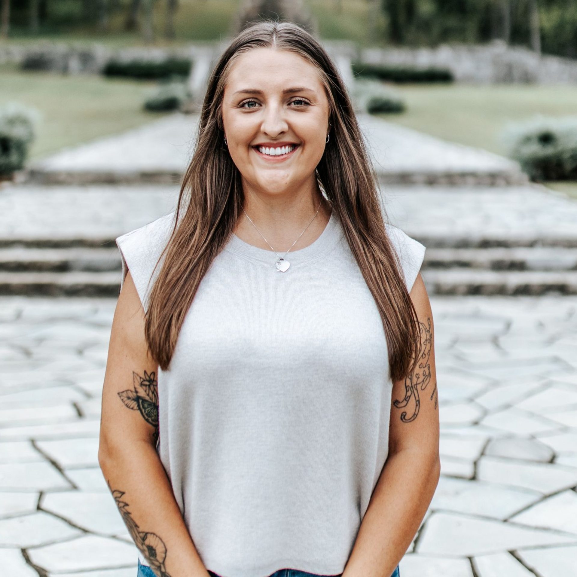 Woman smiling, standing outdoors. She wears a light top, necklace. Arms have tattoos, background with stone steps.