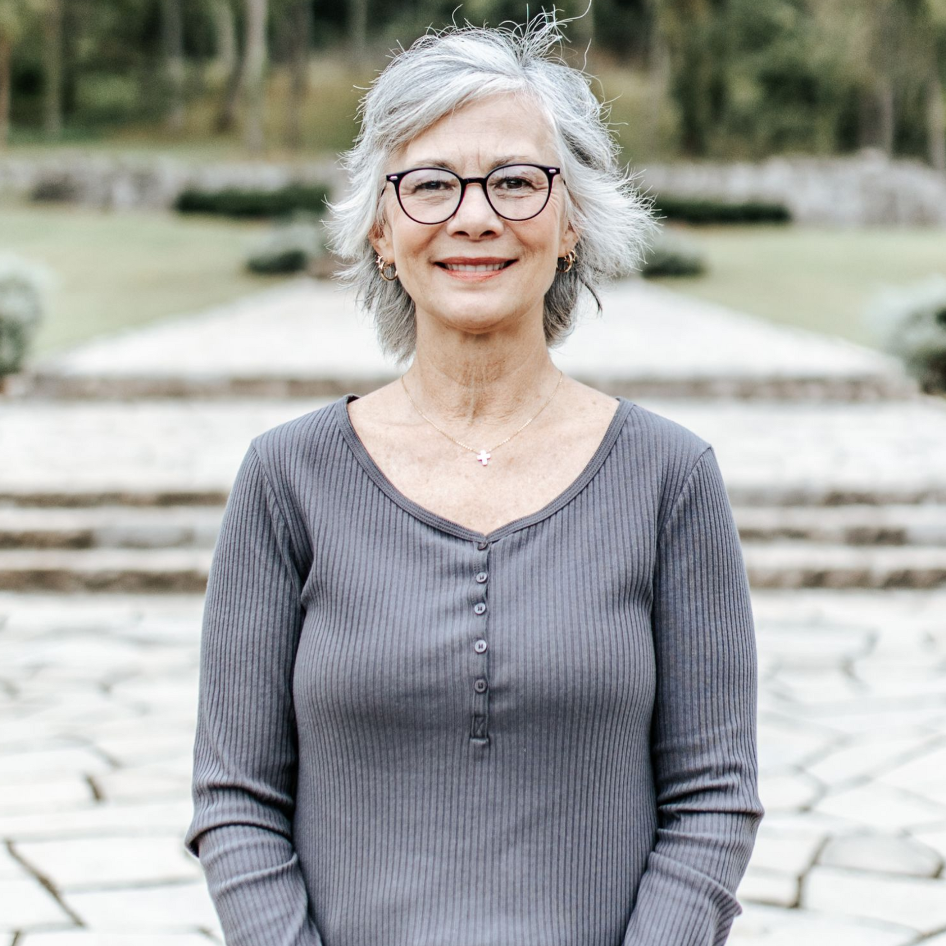 Woman in glasses and gray top smiles outdoors, with a stone background.