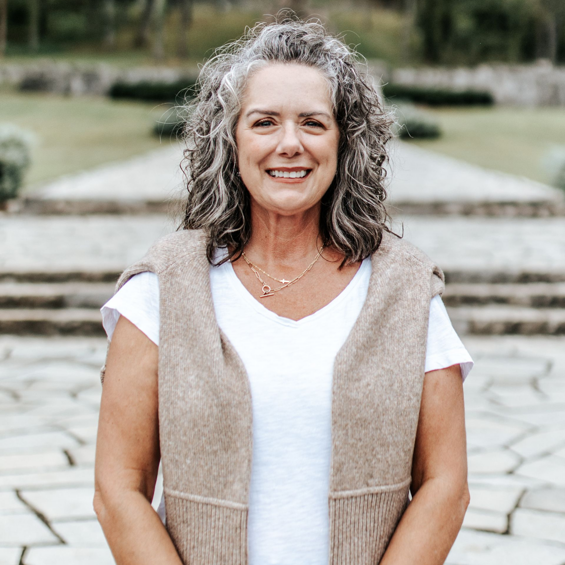 Woman with curly gray hair smiles, wearing a white shirt and beige sweater vest, outdoors near stone steps.