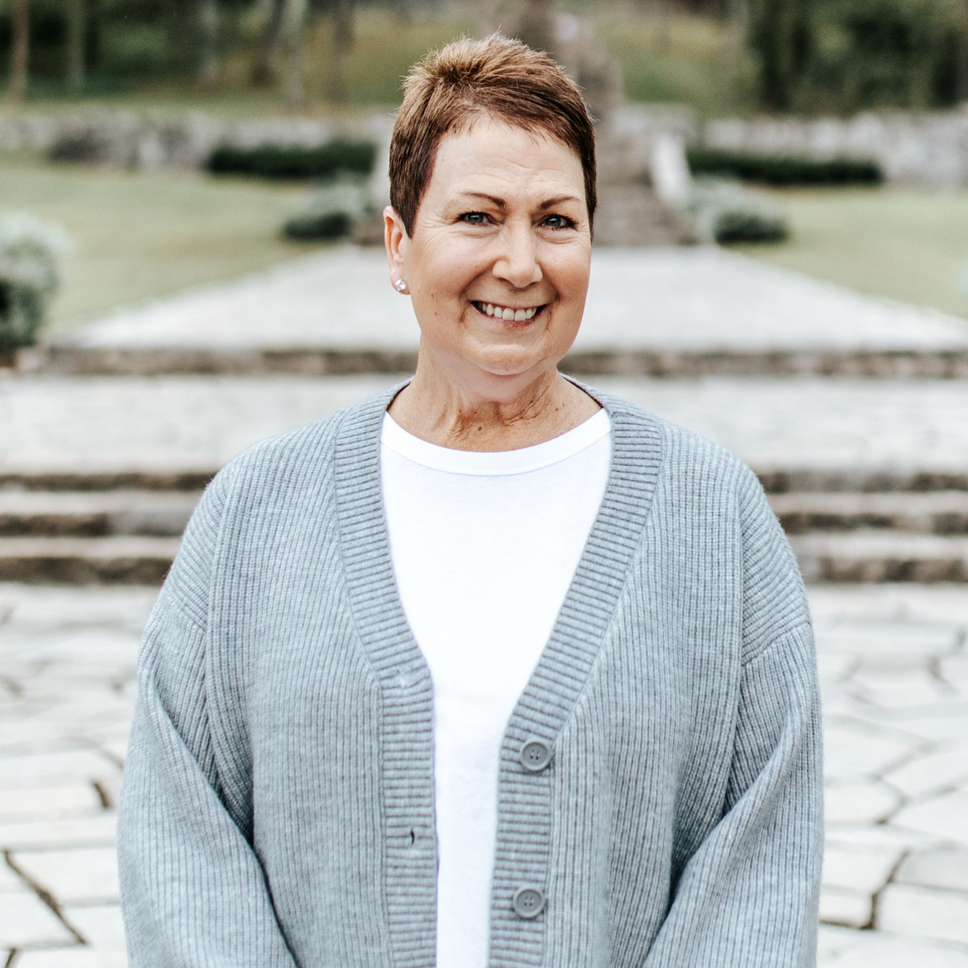 Woman with short brown hair smiles, wearing a gray cardigan over a white shirt, outdoors.