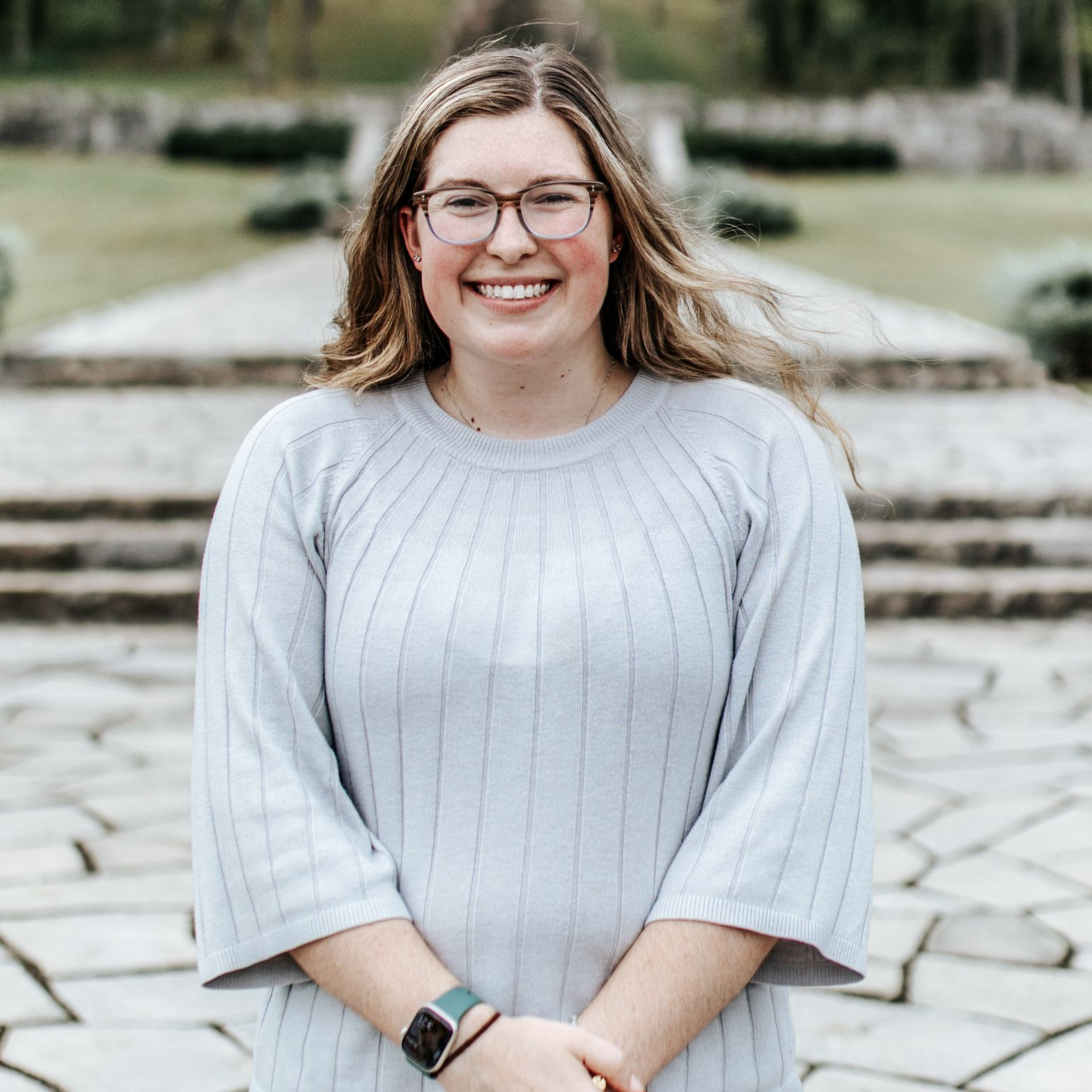 Woman with glasses smiles outdoors, wearing a light blue sweater.