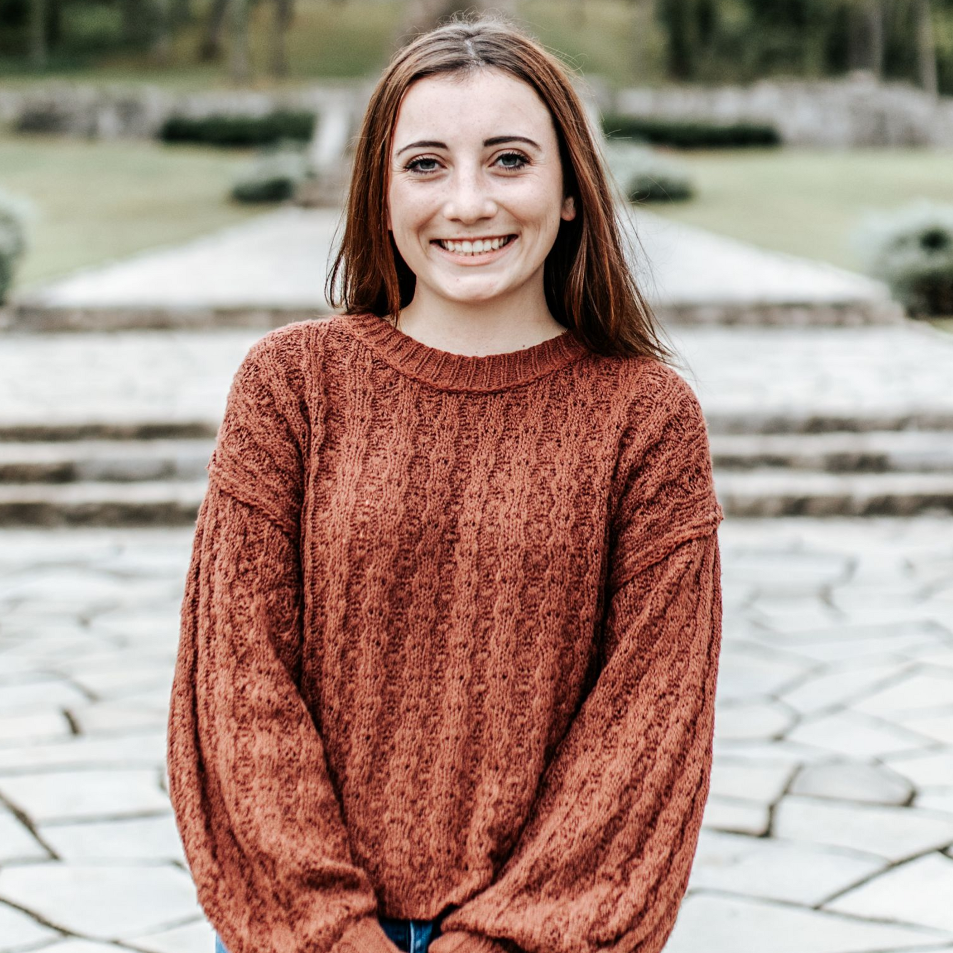 Woman in a burnt orange sweater smiles at the camera in front of a stone walkway.
