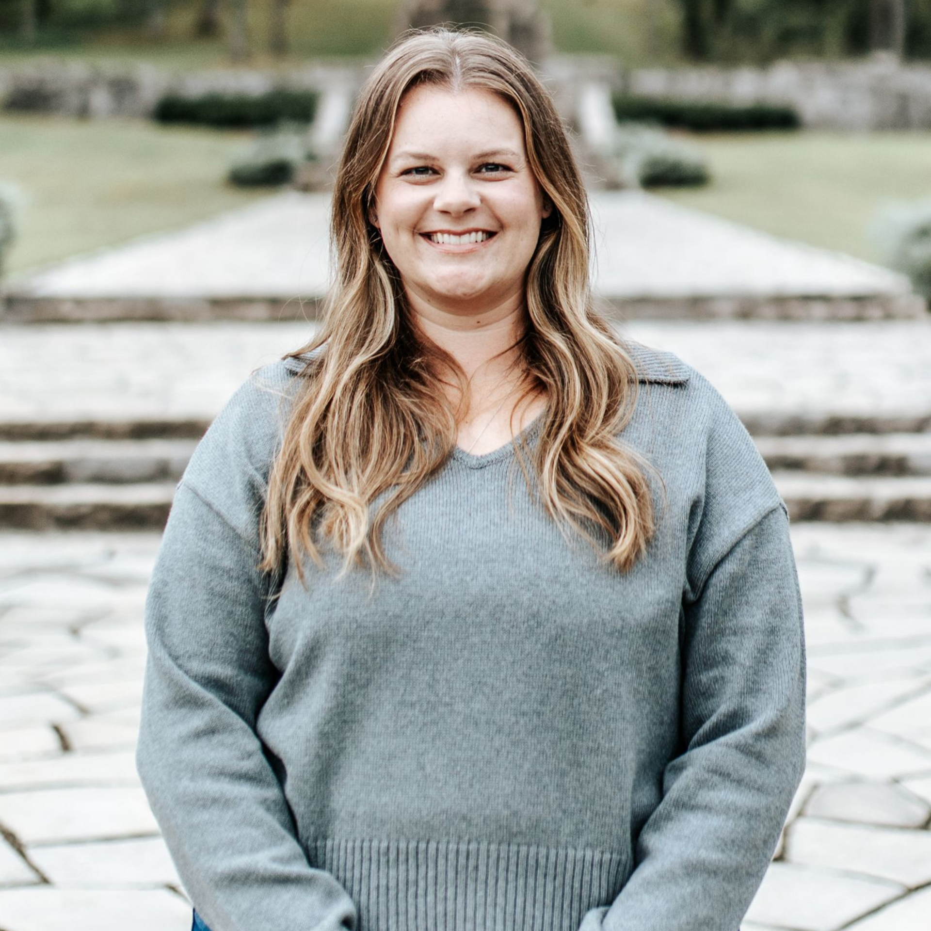Woman with long hair smiles, wearing a blue sweater, standing outdoors. Stone steps and a structure are in the background.