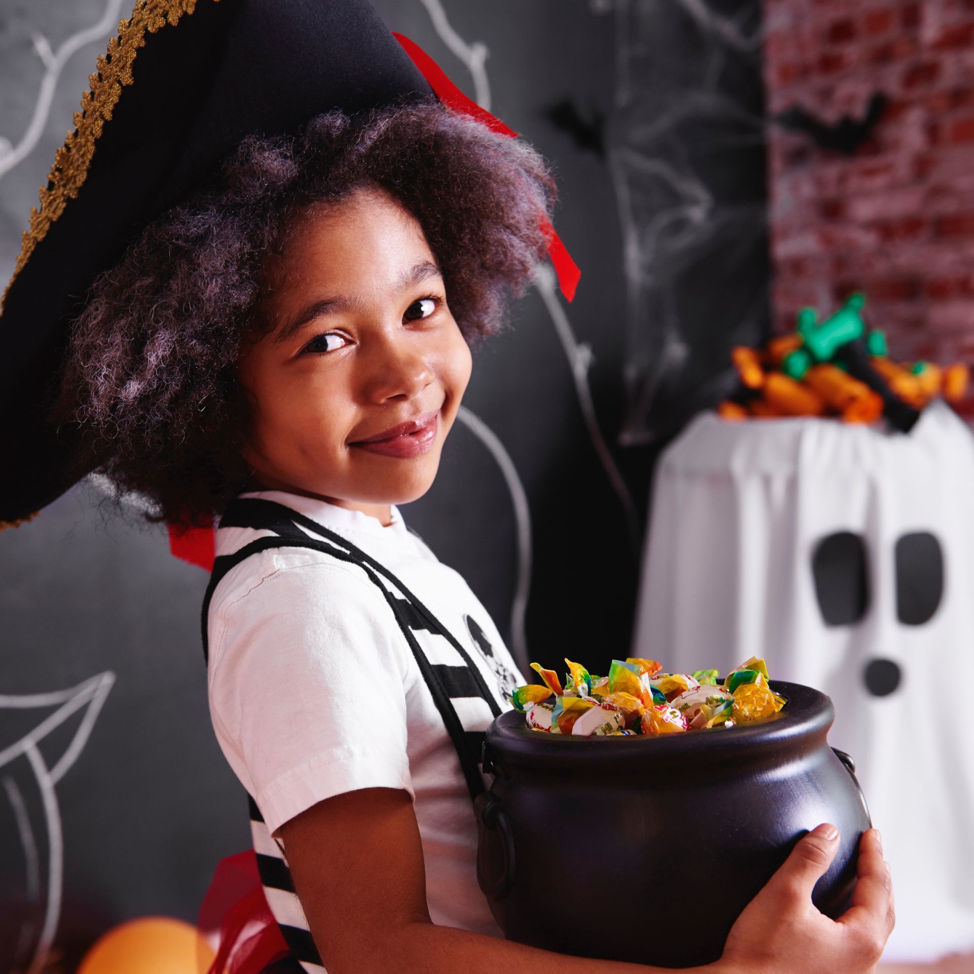 A little girl in a pirate hat is holding a cauldron full of candy