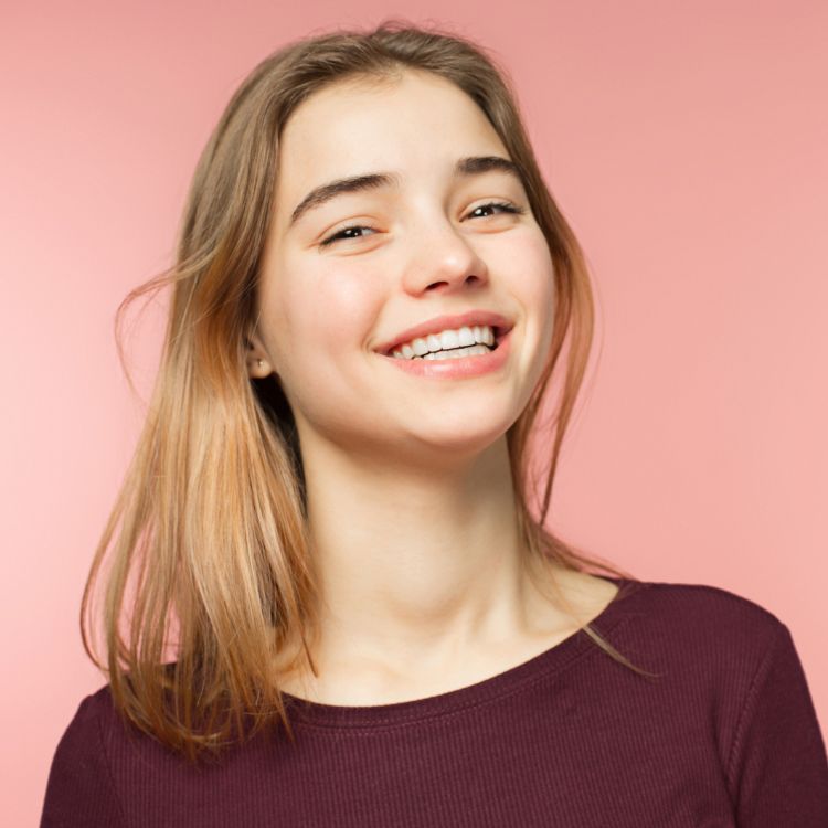 A young woman in a purple shirt is smiling on a pink background.