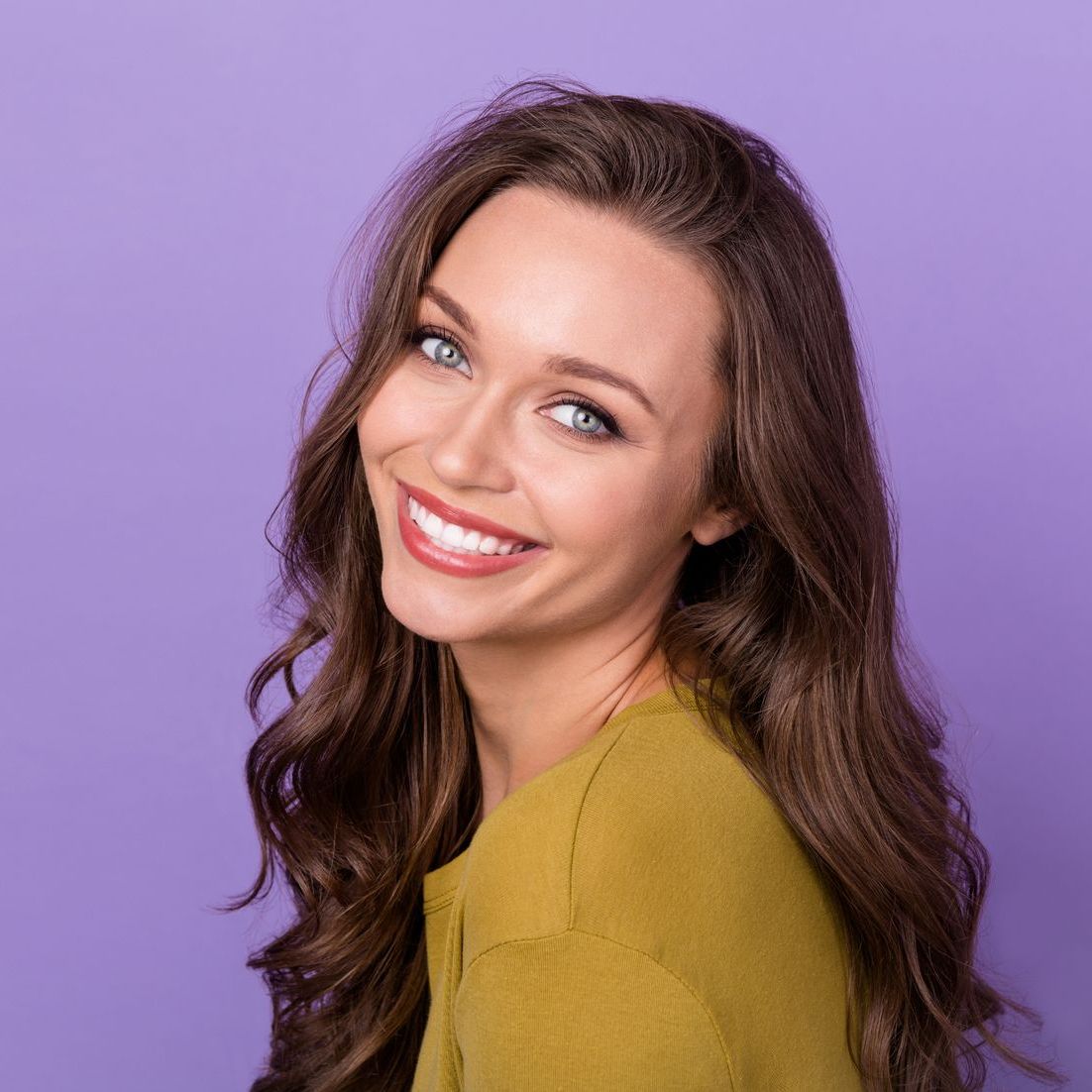 A woman with long hair is smiling and wearing a yellow sweater on a purple background.