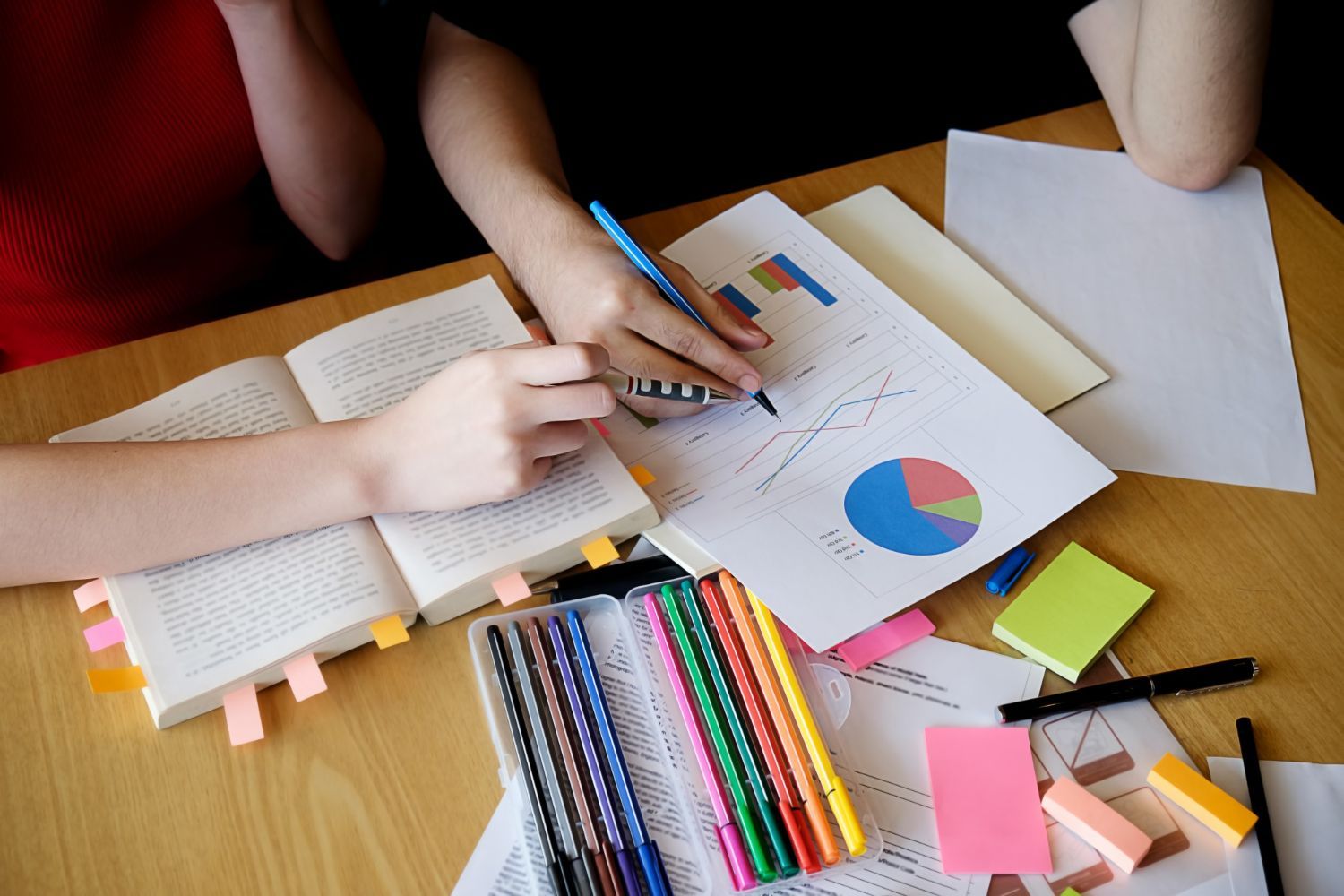 Hands writing on papers with a pie chart and open book, with pens and sticky notes on a wooden desk.