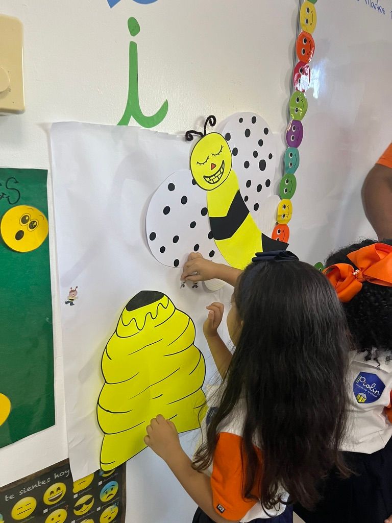 Children placing cutouts of a bee and beehive on paper, in a classroom.