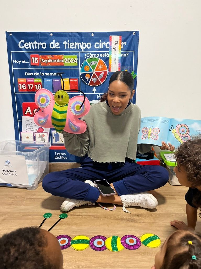 Woman with butterfly puppets, teaching children about a caterpillar on the floor, in front of a colorful poster.