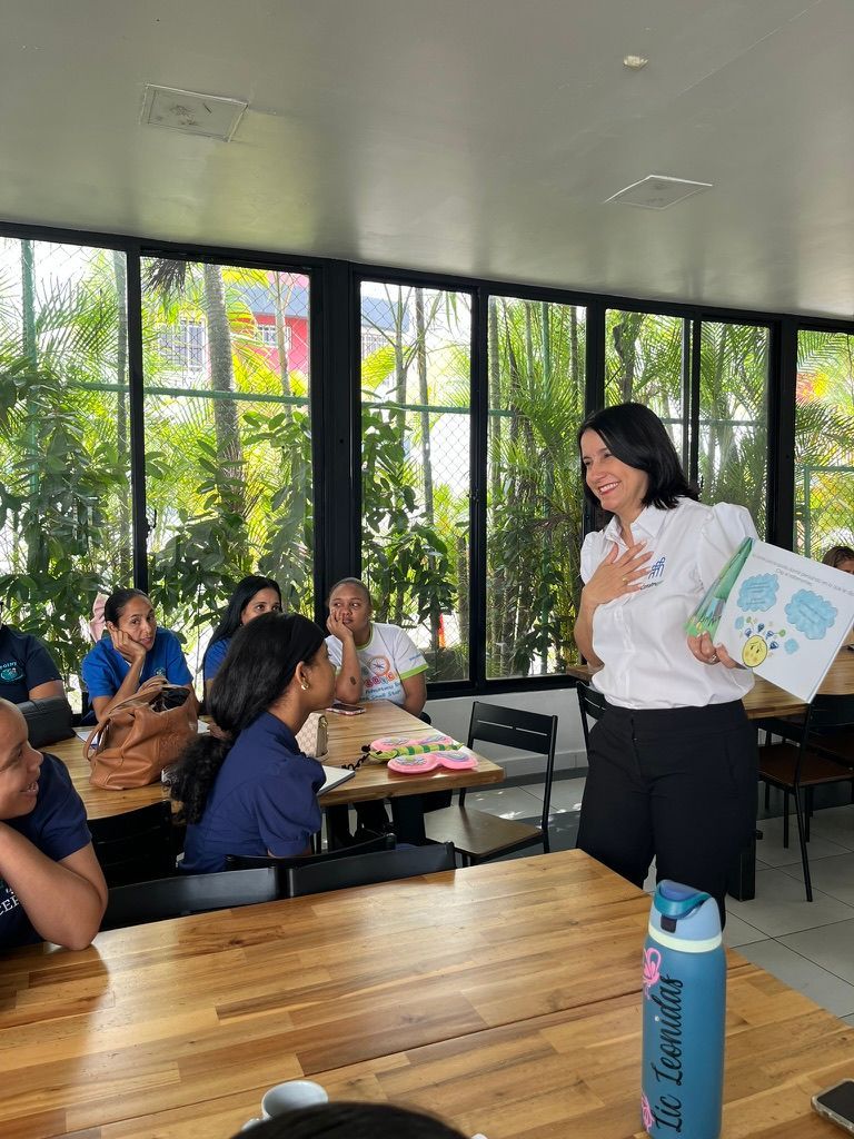 Woman reading a book to a group. People seated at a table, indoors with large windows.