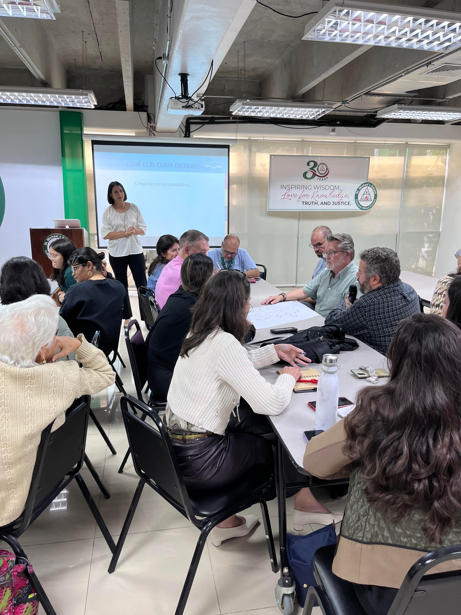 A woman presenting to a group seated around a table, whiteboard in background.