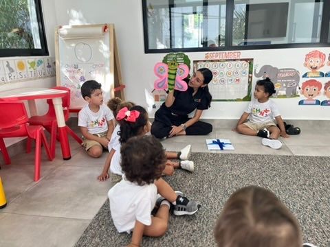 Teacher showing pink shapes to children seated on the floor in a classroom.