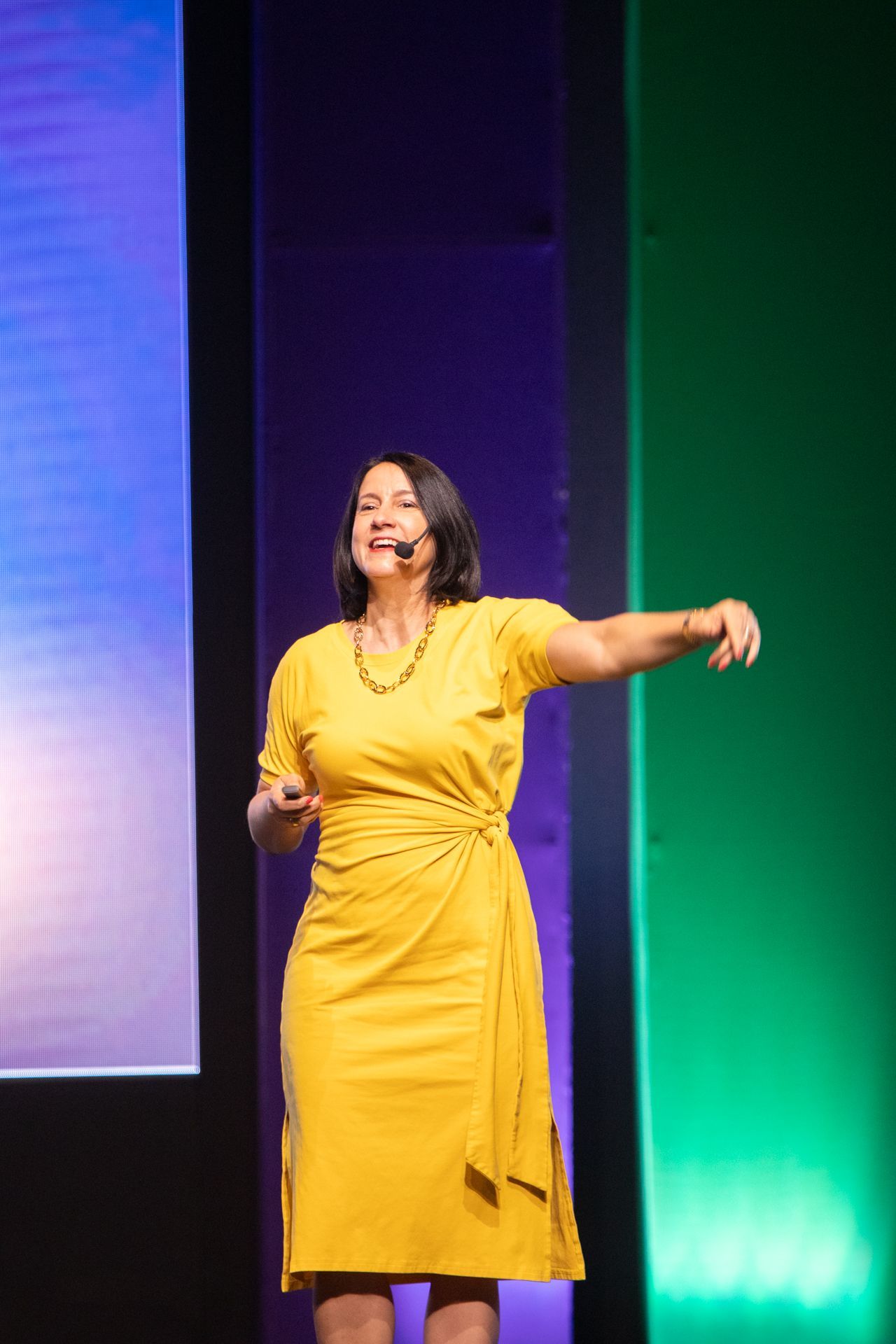 Woman in yellow dress gestures on stage, smiling; green and purple background.