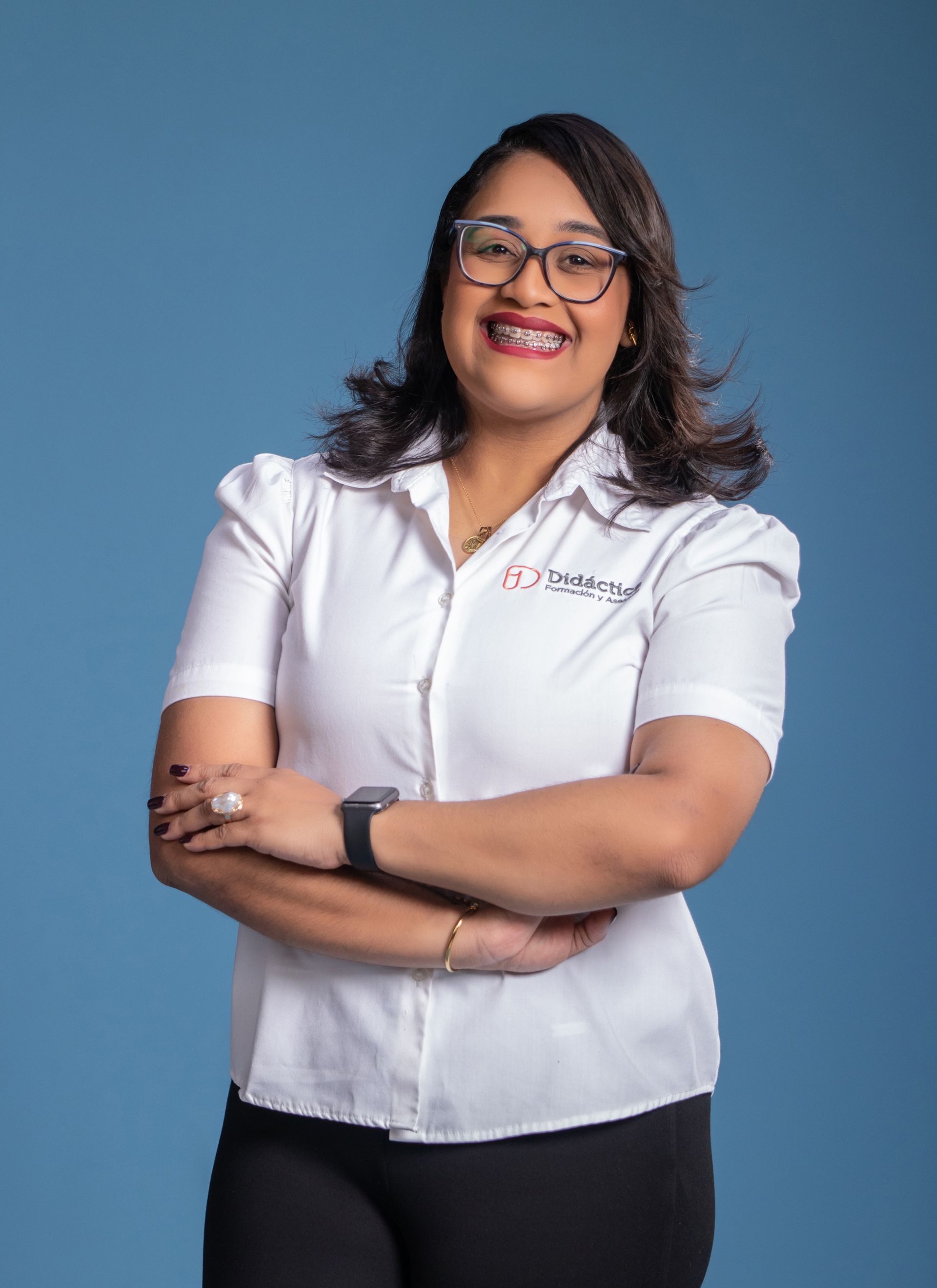 Woman with glasses and braces in a white shirt with logo, arms crossed, smiling. Blue background.