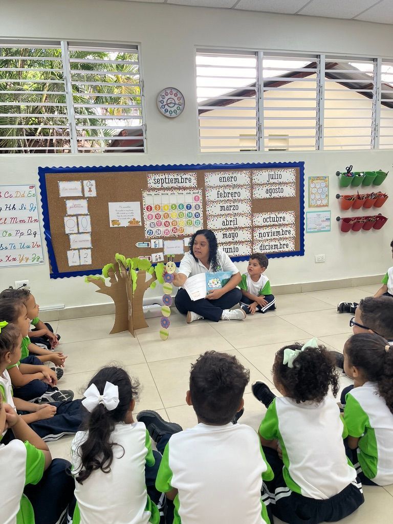 Teacher reading a book to a group of children in a classroom with a tree prop.