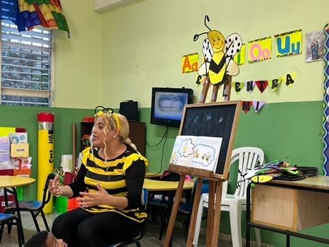 A woman in a bee costume reads a book in a classroom decorated with bee-themed visuals.
