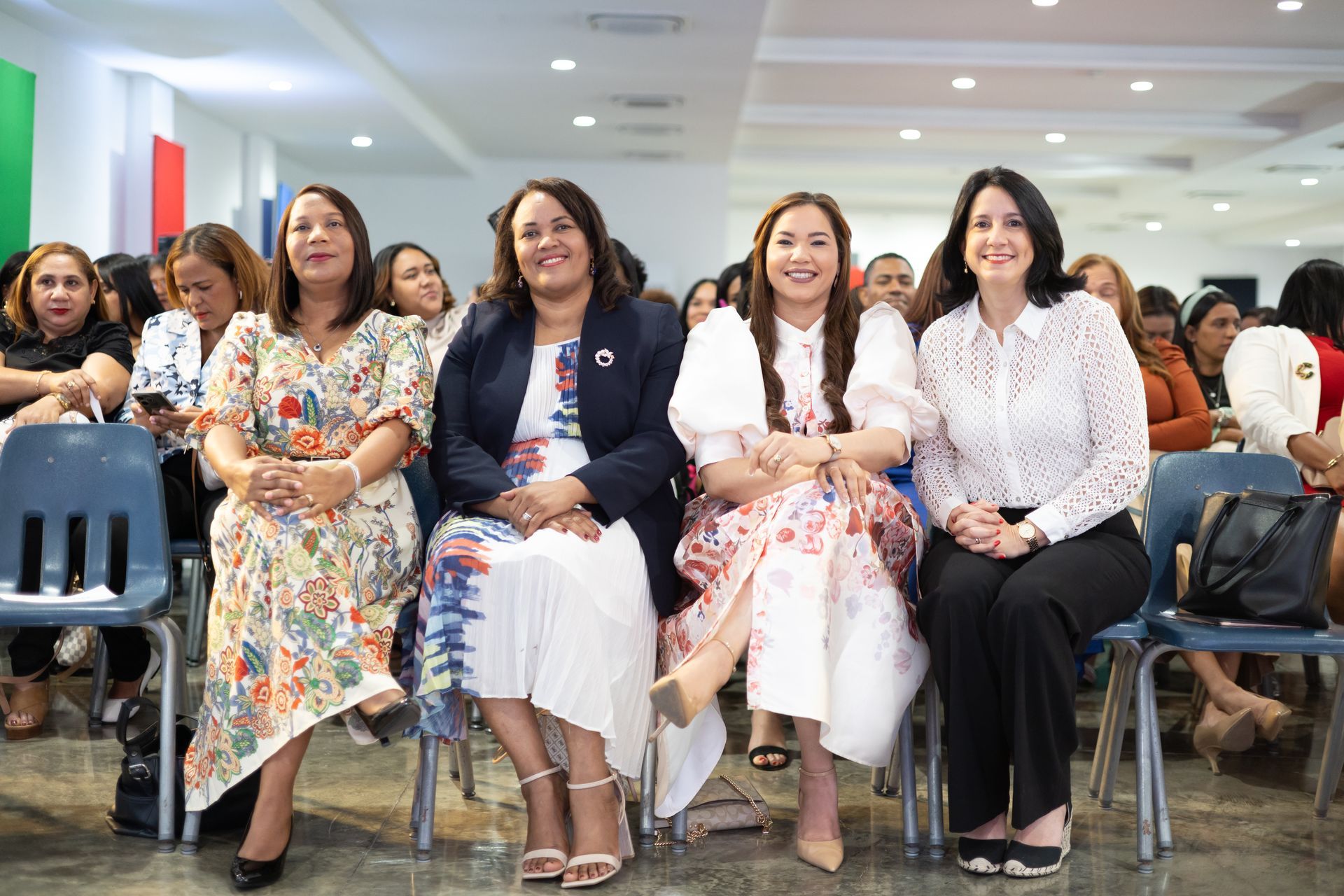 Four women seated, smiling, in a brightly lit room. Two are wearing floral dresses, two are wearing blazers.