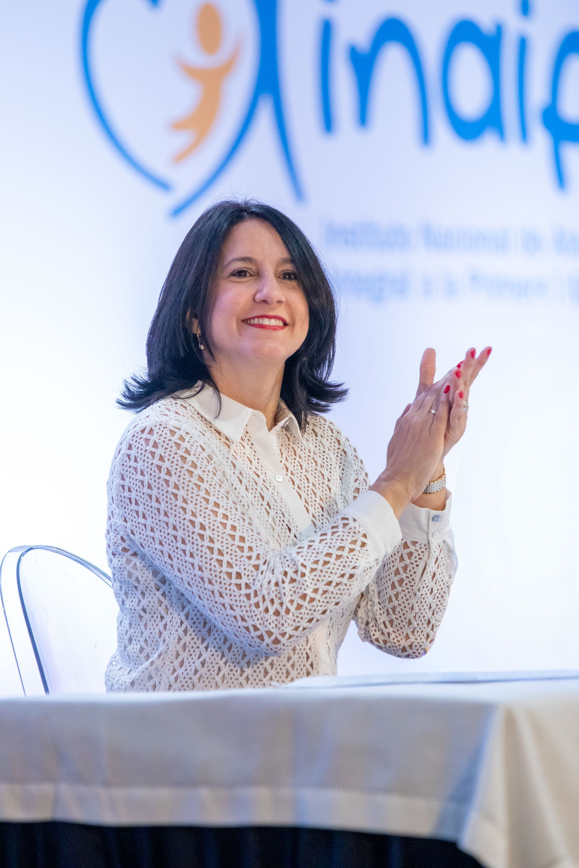 Woman in white shirt clapping at a table, smiling at an event with a logo in the background.