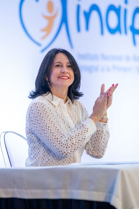 Woman in white shirt clapping at a table, smiling at an event with a logo in the background.