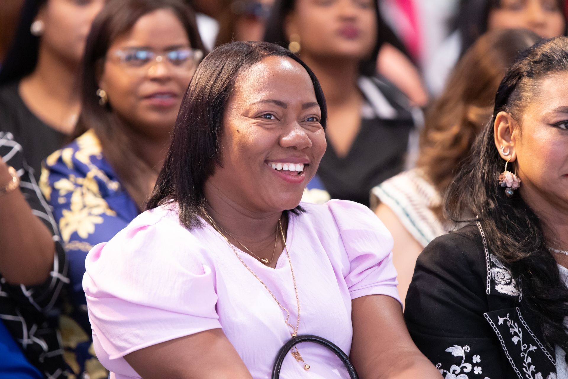 Woman in pink top smiles broadly, seated among other attendees.