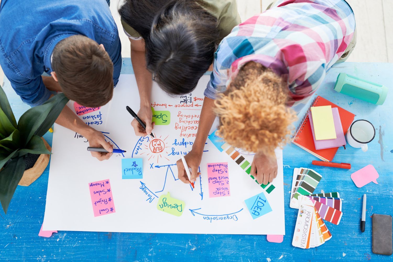 Three people leaning over a table, collaboratively drawing and writing on a large sheet of paper with colorful sticky notes.
