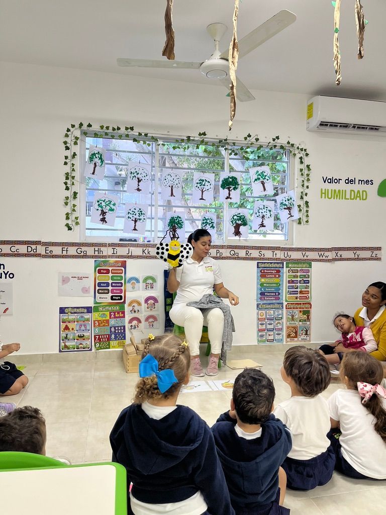 A teacher in a classroom holds a puppet, children seated in front.