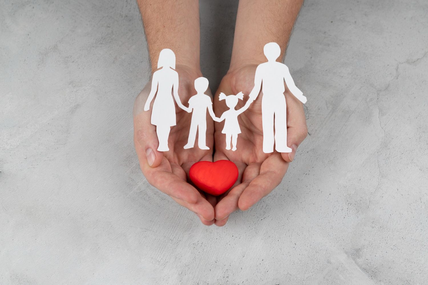 Hands holding a paper family cutout above a red heart on a gray surface.