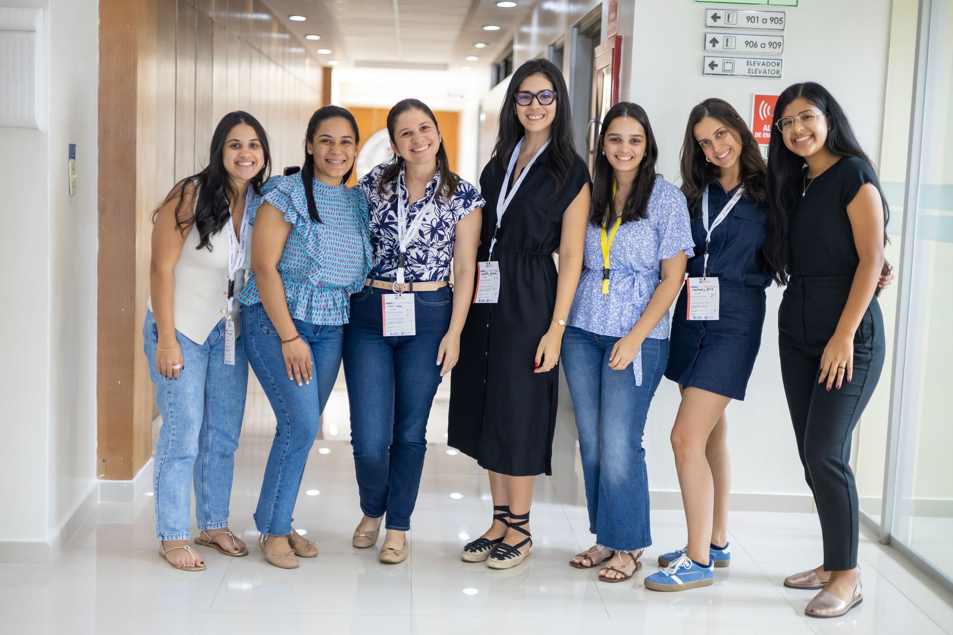Seven women in a well-lit hallway smile at the camera. They wear casual clothes and lanyards.