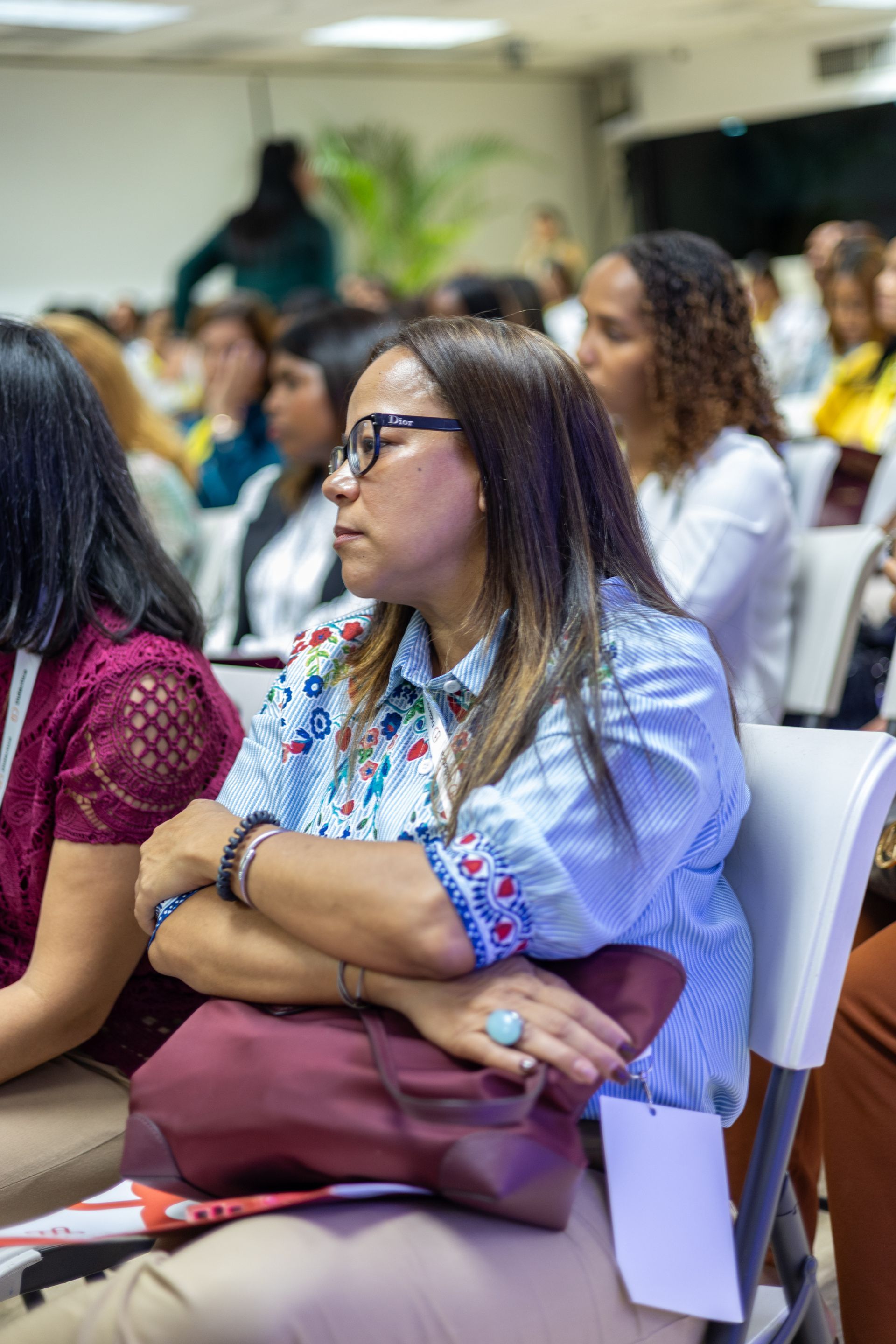 Woman in blue shirt with embroidered details, seated in an audience, arms crossed, looking intently.