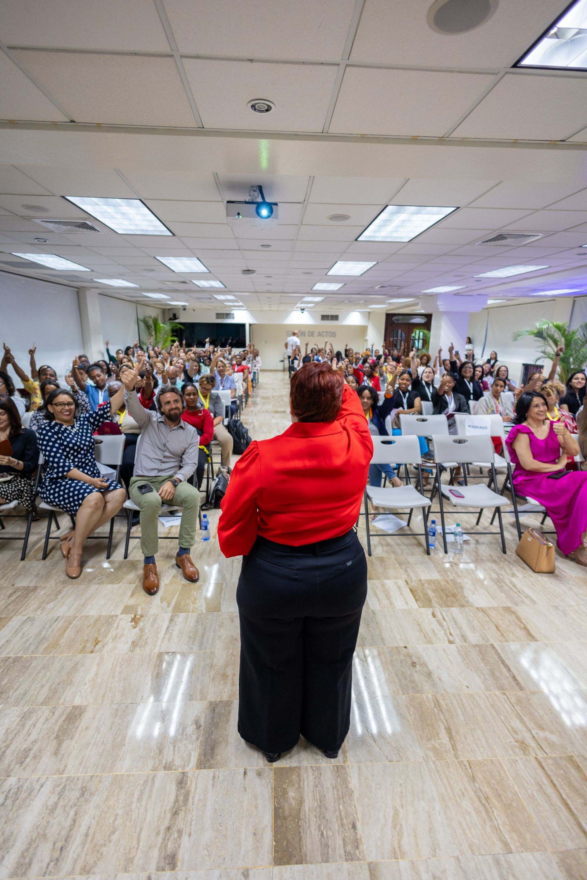 Woman in red shirt addresses a large audience in a bright hall, many people with hands raised.