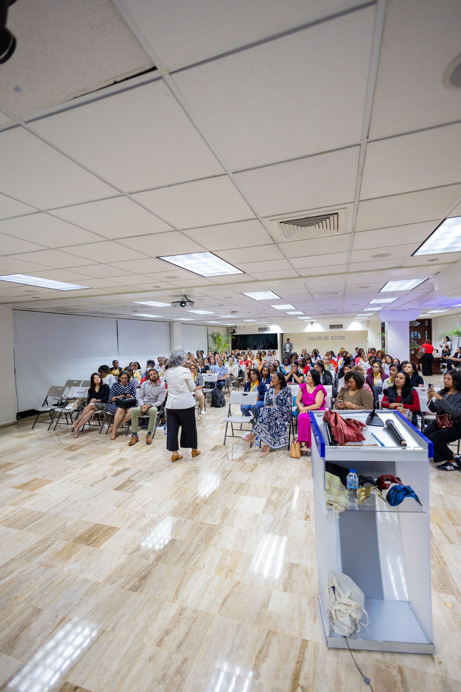 Audience at a presentation, speaker at podium, white room, overhead lighting.