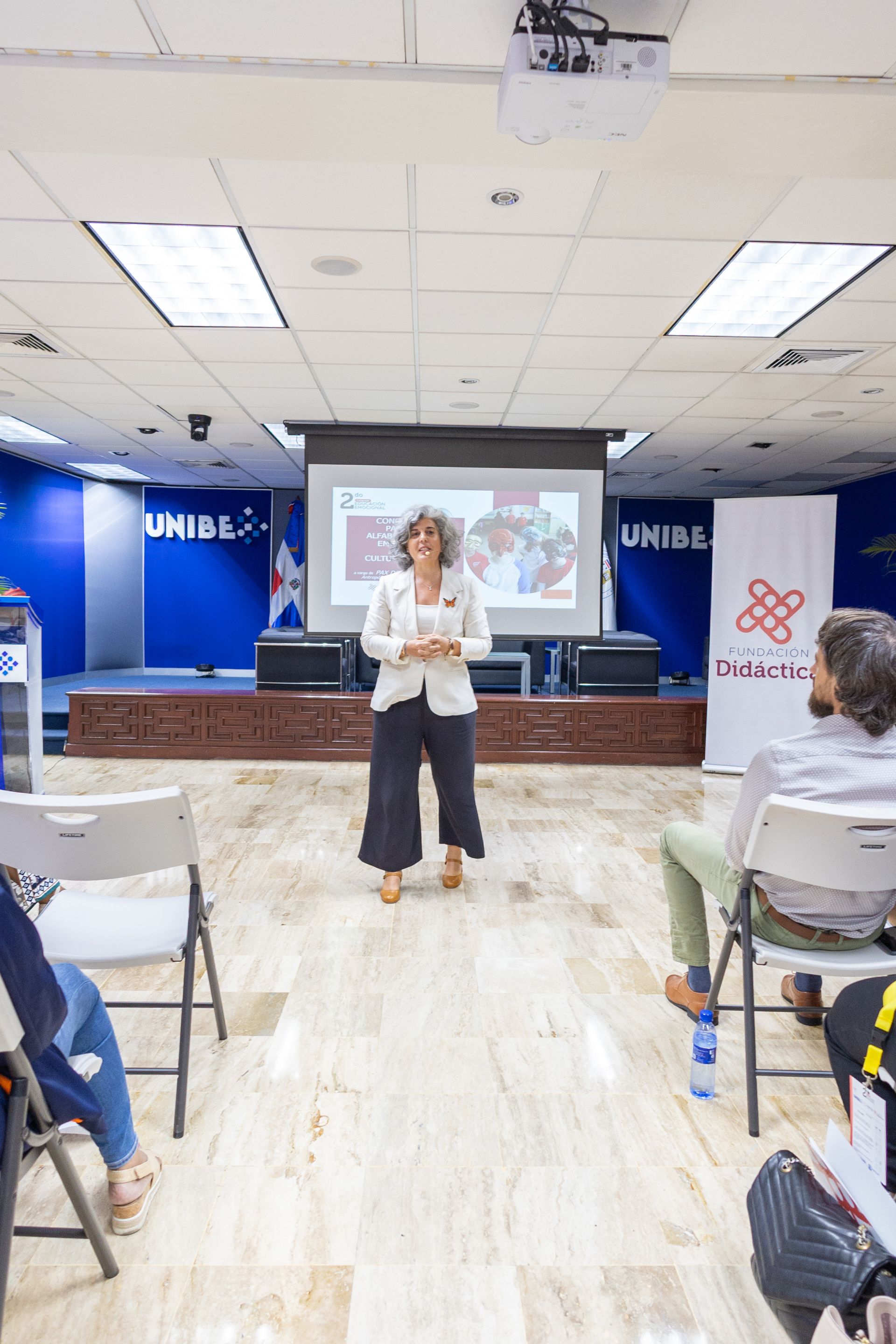 Woman presenting in a room with a projector screen. Audience in chairs. Blue and white decor.