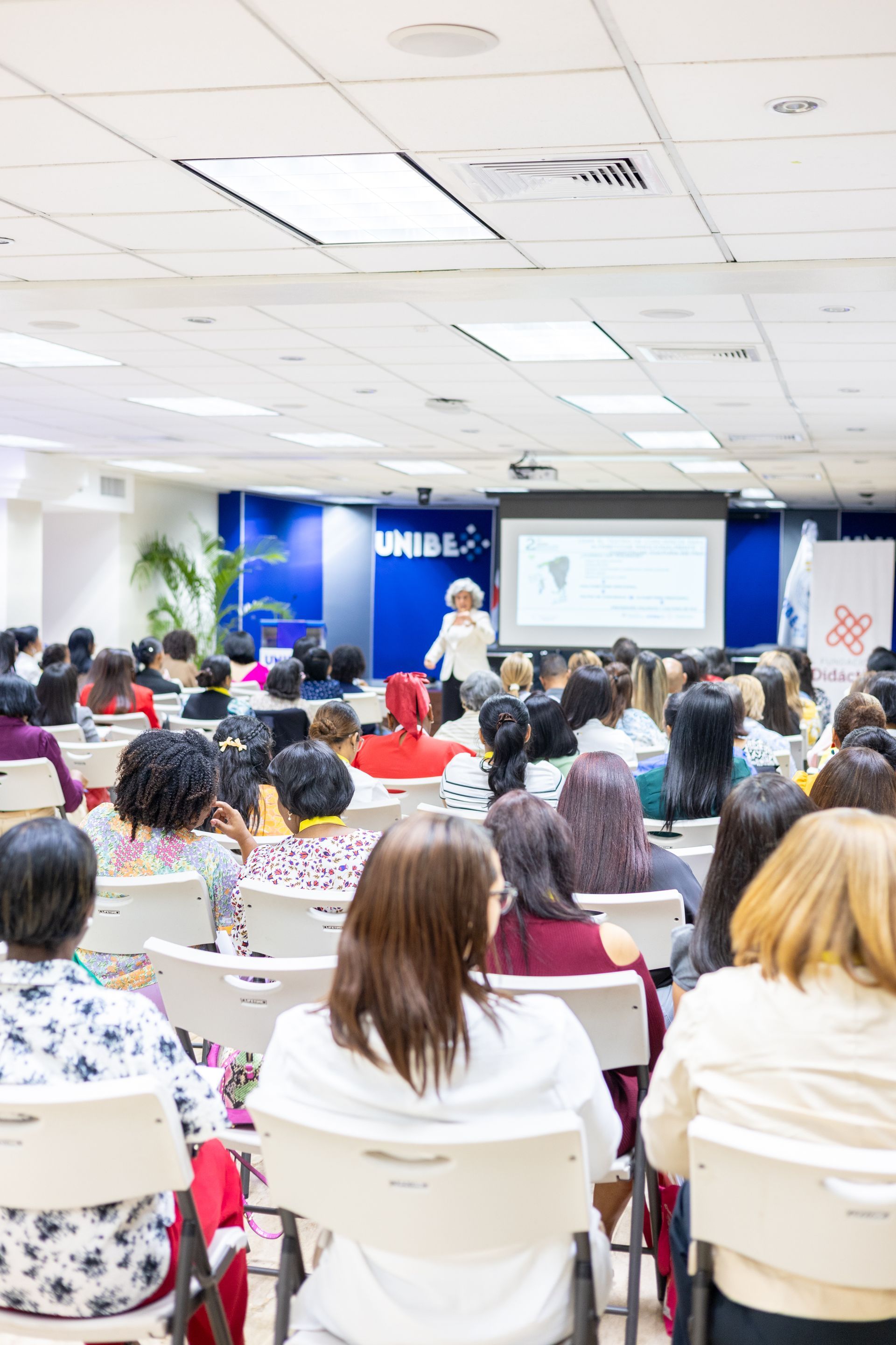 Audience seated in a conference room; a presenter stands near a screen displaying information.