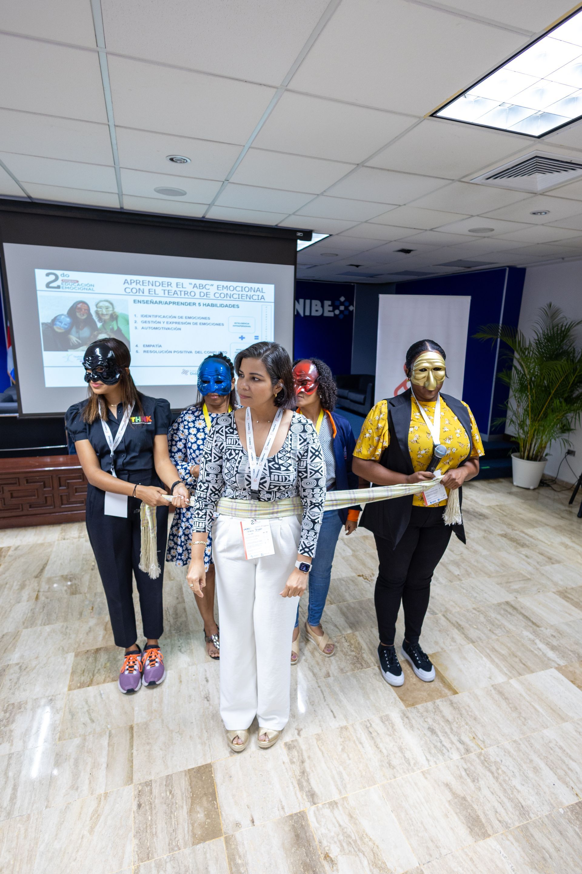 Five people in superhero masks, holding rope, stand in a room. A presentation screen is in the background.