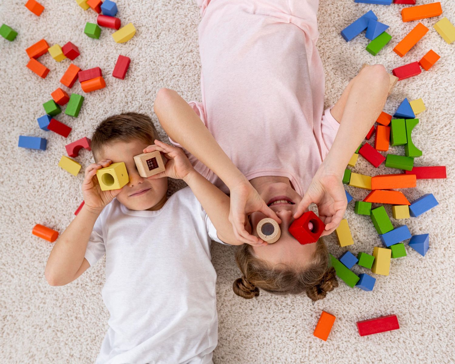 Two children lie on a carpet with blocks, holding them over their eyes.