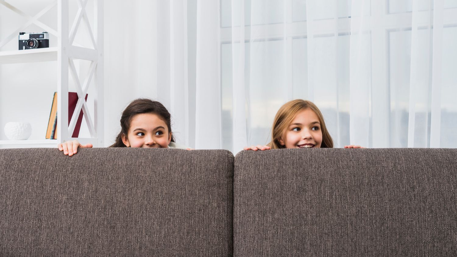 Two children, with brown and blonde hair, peeking over a brown couch, smiling. Interior setting.
