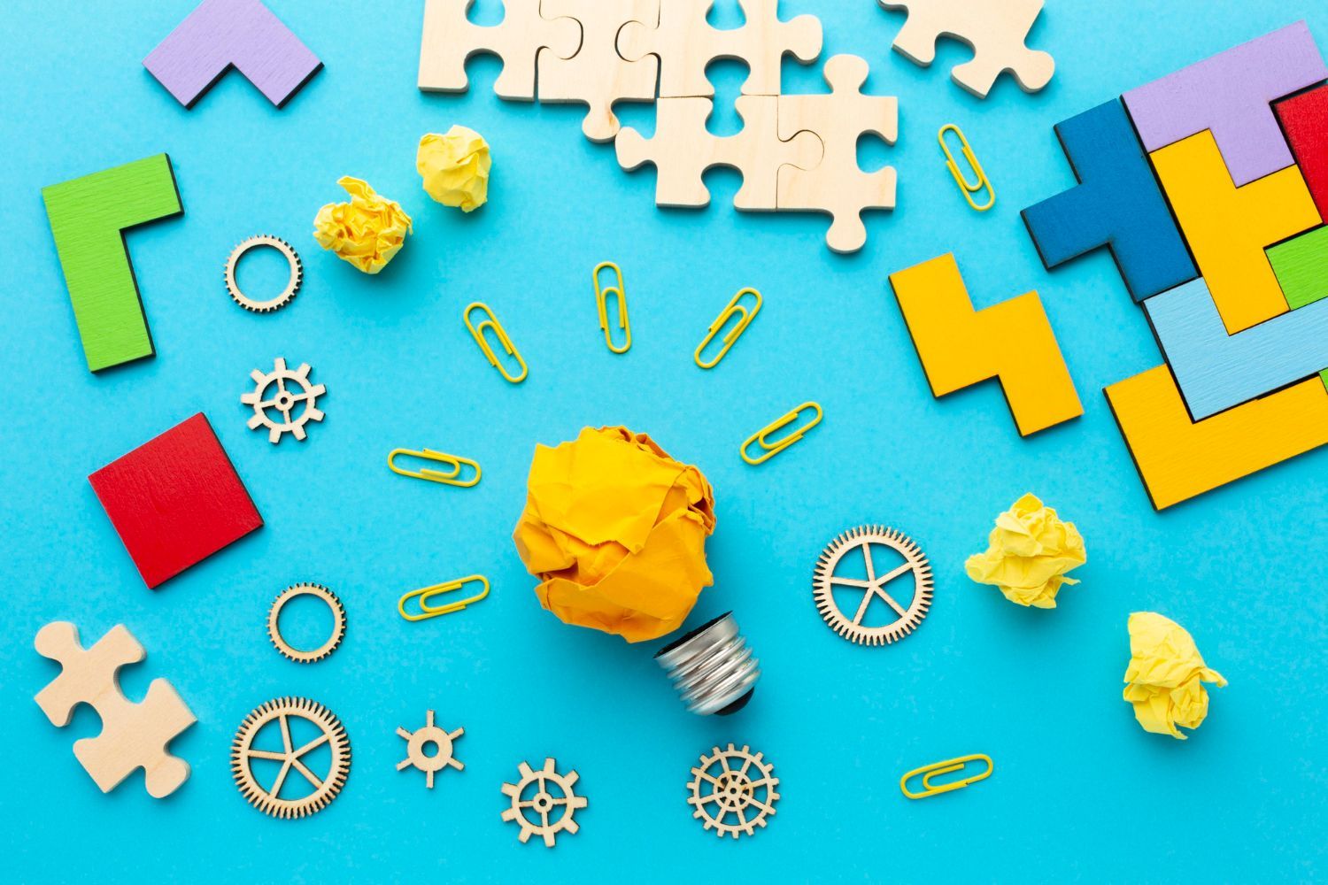 Colorful jigsaw puzzles, gears, paper clips, and crumpled paper arranged on a blue background around a lightbulb made of crumpled paper.