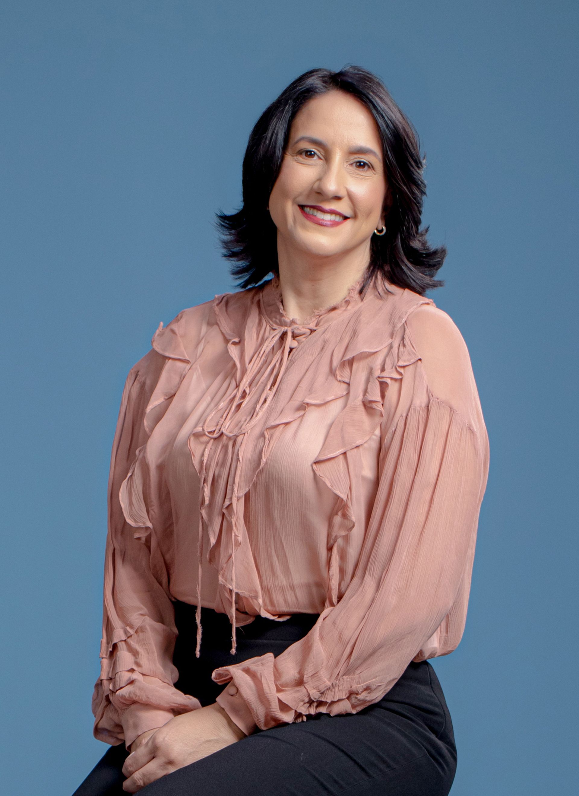 Woman in a pink blouse smiles, seated, against a blue backdrop.